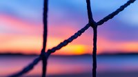 Dramatic sunset sky with vibrant orange and purple colors viewed through silhouetted rope or net lines creating geometric shapes, peaceful seascape horizon background.
