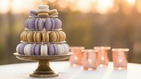 Elegant macaron tower on golden stand with pastel purple and beige French cookies, soft bokeh background perfect for bakery displays and dessert presentations.