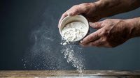 Chef pouring white flour from bowl onto wooden surface, creating dust cloud. Baking and cooking preparation.