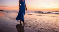 Woman in flowing blue dress walking on beach at sunset with golden sky and ocean waves reflecting warm light creating peaceful seaside moment for travel and lifestyle content.