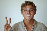 Young Caucasian male with curly hair showing peace sign gesture while smiling at camera, wearing casual beige button-down shirt against light background.