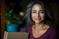 Confident middle-aged Hispanic woman with natural gray hair smiling while holding cardboard box, wearing burgundy top against blurred background with houseplant.