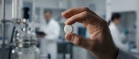 Scientist holding white pill or tablet in laboratory setting with blurred colleagues and equipment in background, representing pharmaceutical research and development.