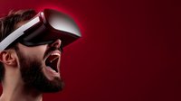 Bearded man wearing virtual reality headset expressing excitement and amazement against dark red background, showing immersive technology experience.