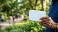 Man holding blank white card outdoors in park with green foliage background, suitable for advertising, marketing messages, business promotions, and communication concepts.