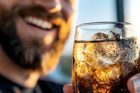 Smiling bearded man enjoying whiskey or cola drink with ice cubes in glass during golden hour sunset celebration.