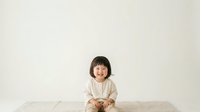 Young child with dark hair and fair complexion, wearing a cream-colored long-sleeve shirt, sitting on a white bed against a neutral background. Suitable for parenting, childcare, and family lifestyle content.