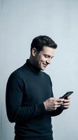 Young man with dark hair and light skin tone, wearing a charcoal turtleneck sweater, smiling while using a smartphone against a neutral white background. Suitable for technology, communication, and lifestyle applications.