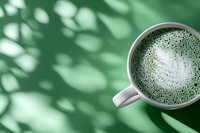 Hot matcha green tea latte with foam art pattern in white ceramic cup on green background with natural shadow overlay from leaves.