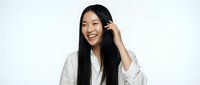 Young Asian woman with long black hair wearing white shirt, touching hair and smiling at camera against white background for portrait and lifestyle use.