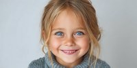 Close-up portrait of smiling young girl with blue eyes and blonde hair wearing gray sweater against neutral background.