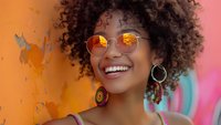 Young African American woman with natural curly hair and round glasses smiling against colorful background, expressing joy and confidence in casual summer style.