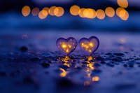 Two glowing heart-shaped objects with warm golden lights inside rest on wet pavement during blue hour, creating romantic bokeh reflections for love concepts.