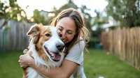 Young woman with long wavy hair wearing white t-shirt hugging and sharing joyful moment with Australian shepherd dog in backyard garden during golden hour sunset.