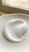 Creamy white cosmetic product swirled on neutral plate near window, showcasing skincare or beauty formulation texture for product photography and cosmetics marketing.