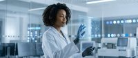 Female scientist of African descent wearing white lab coat and blue nitrile gloves, working in modern laboratory with advanced equipment and monitors in background, conducting research or analysis.