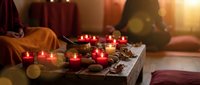Cozy meditation space with burning candles, crystals and spiritual items on wooden tray. Person in orange clothing sits peacefully in warm candlelit room.