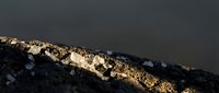 Mountain ridge with snow patches illuminated by golden sunlight against dramatic dark sky creating striking contrast for outdoor adventure content.