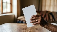 Senior man with dark skin tone holding blank white book or magazine on wooden table indoors, suitable for publishing, education, and marketing materials.
