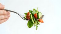 Fresh mixed greens salad with cherry tomatoes and cucumber on fork held by hand against white background for healthy eating concepts.