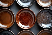 Empty ceramic plates in earthy brown and black tones with textured surfaces arranged in rows on dark background, creating minimalist dining composition.