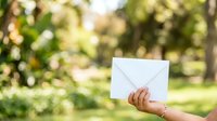 Woman holding blank white envelope outdoors in sunny garden setting with green foliage background, suitable for mail, communication, and correspondence concepts.