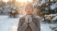 Young woman in winter coat and knit hat enjoying peaceful moment in snowy forest with warm sunlight filtering through trees during golden hour snowfall.