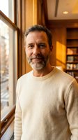 Mature Caucasian man with beard in cream sweater standing by window in warm cozy home interior with natural lighting and wooden shelves in background.