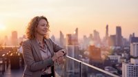 Businesswoman with curly reddish-brown hair wearing professional blazer standing on rooftop overlooking city skyline at sunset, checking smartwatch, urban professional lifestyle.