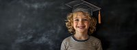 Young caucasian boy with curly blonde hair wearing striped shirt and graduation cap smiles brightly against dark chalkboard background, education concept.