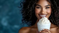 Young African American woman with curly hair smiling while holding a whipped cream dessert against blue background. Perfect for food, beauty, or lifestyle marketing.