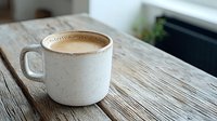 Creamy coffee in white ceramic mug on rustic wooden table surface, morning light creates cozy atmosphere for cafe and lifestyle photography.