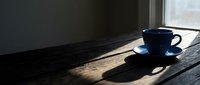 Dark blue ceramic cup and saucer on wooden table by window with natural sunlight casting shadows, minimalist morning coffee scene for lifestyle and beverage content.