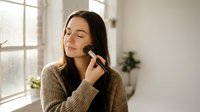 Young woman applying makeup with brush in bright modern interior enjoying morning beauty routine at home with natural window light.