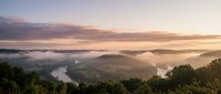 Aerial view of misty river valley at sunrise with forested hills and golden light breaking through clouds, suitable for travel and nature backgrounds.