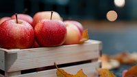 Fresh red apples with water drops in wooden crate, autumn leaves and blurred bokeh background. Natural organic produce for healthy eating and harvest season.