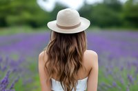 Young woman in straw hat and white dress standing in blooming lavender field, view from behind showing long wavy brown hair against purple flowers.