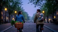 Couple cycling together on cobblestone street at twilight with basket of flowers, romantic evening bike ride through tree-lined urban neighborhood with glowing streetlights.