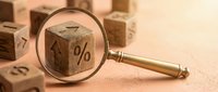 Magnifying glass examining wooden cube with percentage symbol among scattered blocks representing financial analysis and interest rate investigation.
