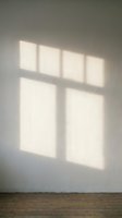 Sunlight streaming through window panes casts geometric shadows on neutral wall, creating minimalist interior composition with wooden floor for architectural and design projects.