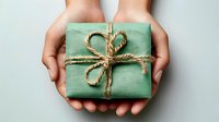 Hands holding small mint green gift box wrapped with natural twine bow against neutral background. Perfect for holiday or celebration concepts.