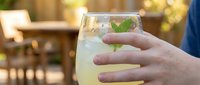 Closeup of hand holding glass of refreshing lemonade with mint leaf and ice cubes outdoors on sunny day, summer beverage concept for advertising refreshments.