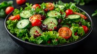 Fresh garden salad with sliced cucumbers, cherry tomatoes, mixed greens and pink peppercorns served on black plate against dark background. Food photography.