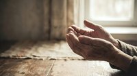 Elderly hands in prayer position on wooden table near window with soft natural light creating peaceful spiritual atmosphere for meditation and faith.