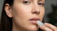Young Caucasian woman applying white lip balm stick to lips for skincare routine and lip care protection in natural indoor lighting.