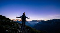 Young Caucasian male hiker stands with arms outstretched on a mountain trail at twilight, embracing freedom under a starry blue sky.