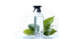 Clear spray bottle with natural cleaning solution surrounded by fresh green leaves and water droplets on white background, eco-friendly household concept.