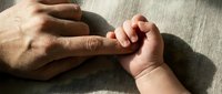 Adult hand gently holding tiny newborn baby hand in tender moment of connection and love on soft neutral fabric background.