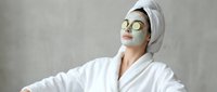Young woman in white bathrobe with cucumber slices on eyes and towel on head relaxing during spa treatment for skincare and wellness routine.