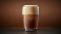 Cold glass of dark craft beer with thick frothy head and condensation drops on brown background, dramatic studio lighting creates appetizing beverage photo.